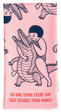 Pink tea towel with illustration of a child on an alligator and text 'Do one thing every day that scares your family' on a white background.